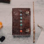 Flat lay of a leather journal with chakra stones, a wand, and mystical elements on a wooden surface.