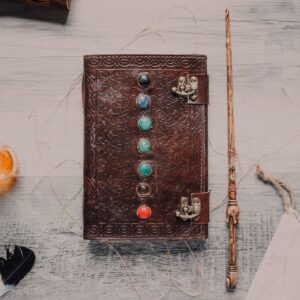 Flat lay of a leather journal with chakra stones, a wand, and mystical elements on a wooden surface.